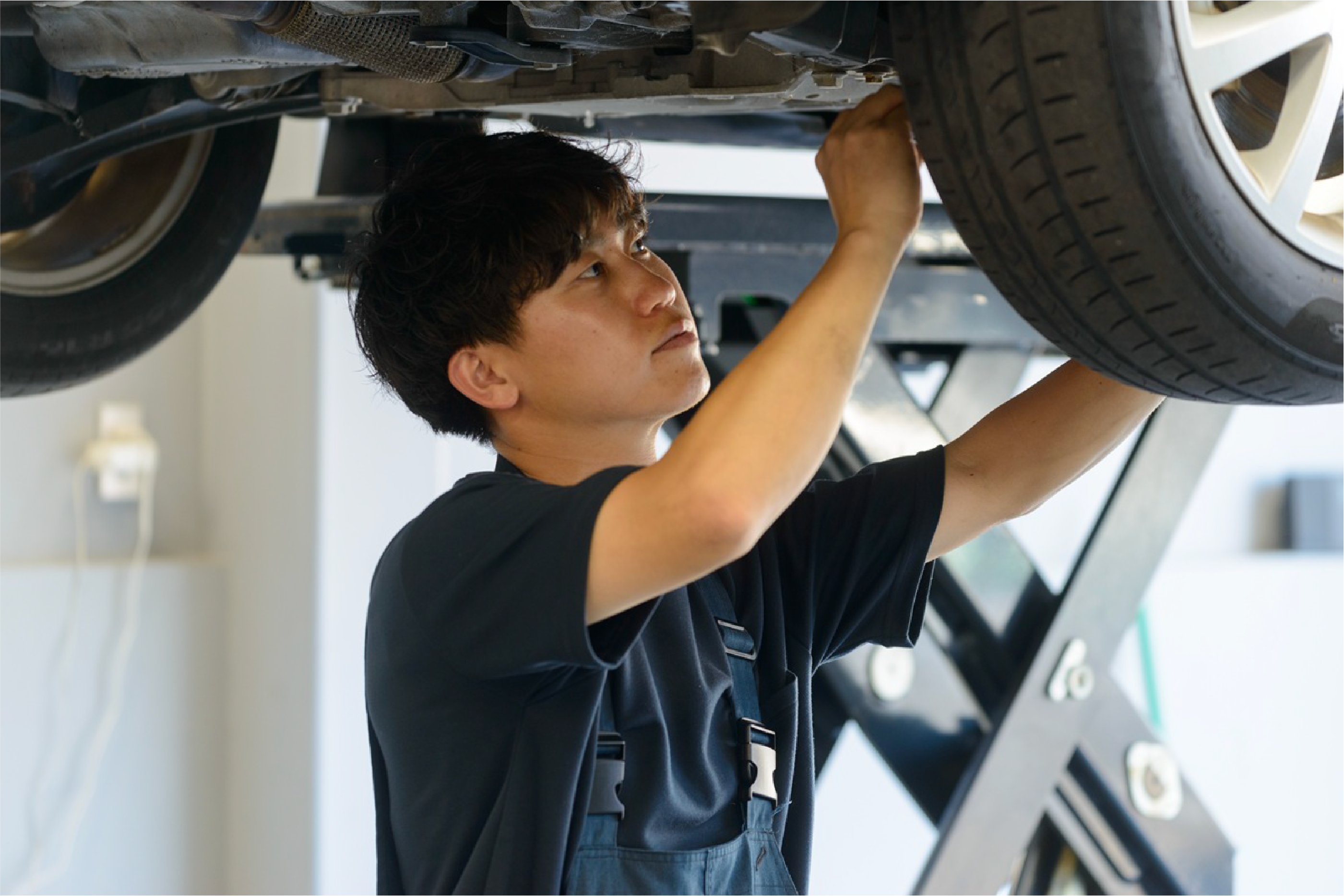 青いツナギを着た男性が、リフトで持ち上げた車の下に入って車の整備している写真