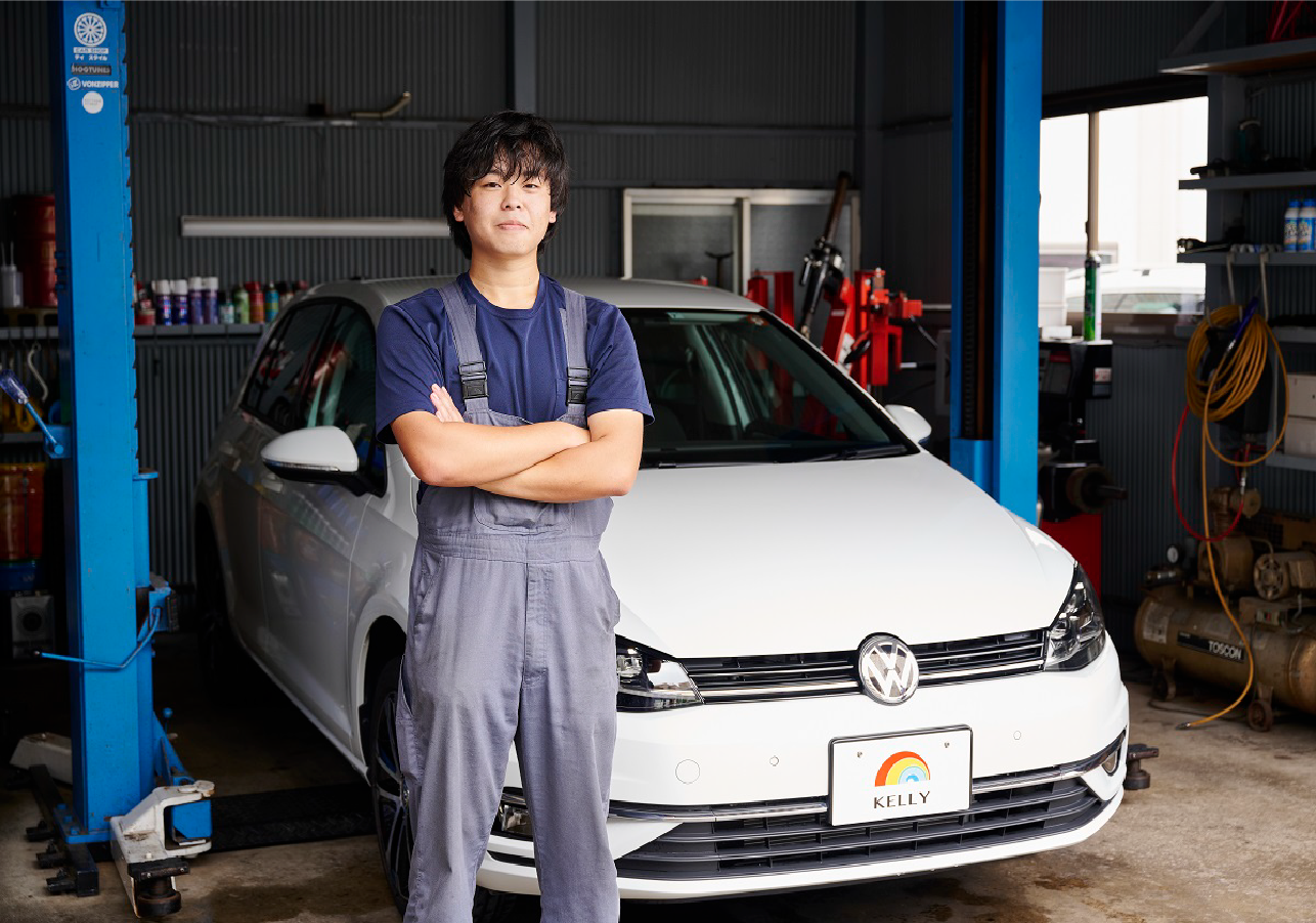 工場内で車の前に立ち、青いツナギを着た男性が腕を組んでいる写真