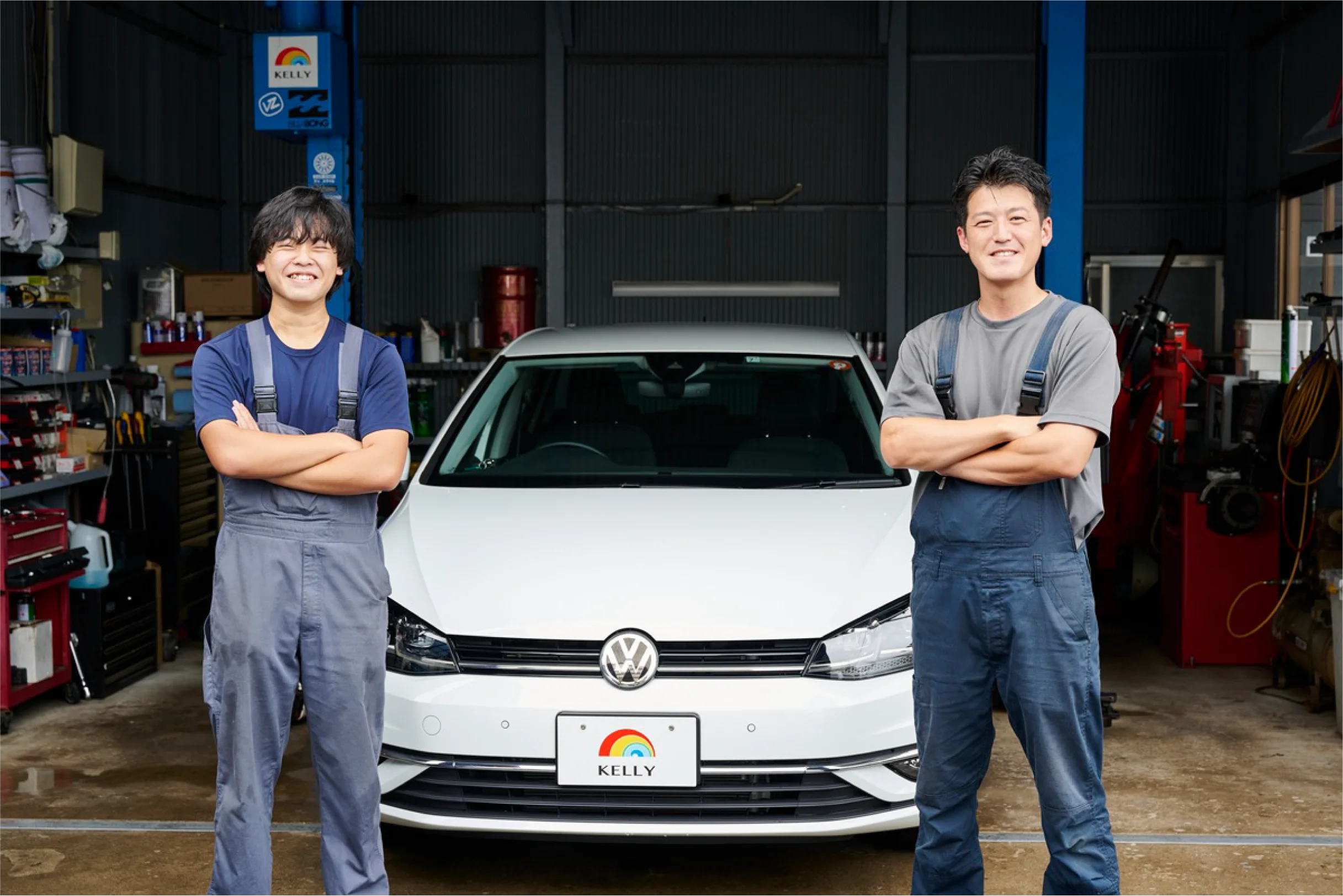 青いツナギを着た整備員の男性二人が車の前で腕を組んでいる画像