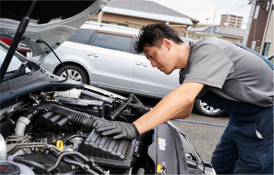 車のボンネットを開け、右手に持ったライトで修理箇所を調査する職員の写真